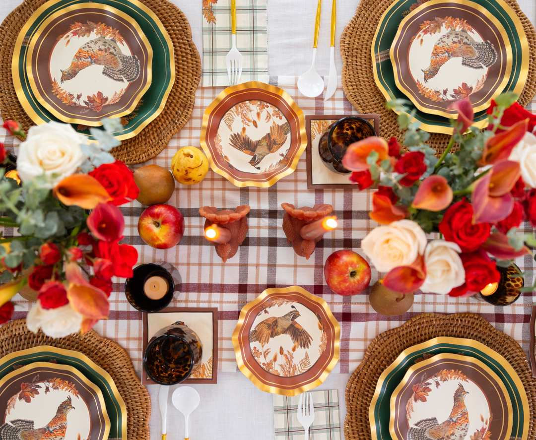 The Woodland Harvest Table Setting by Sophistiplate creates a festive fall-themed tablescape with a plaid tablecloth, floral arrangements, gold-trimmed plates featuring bird designs, apples, pears, candles, and woven placemats.