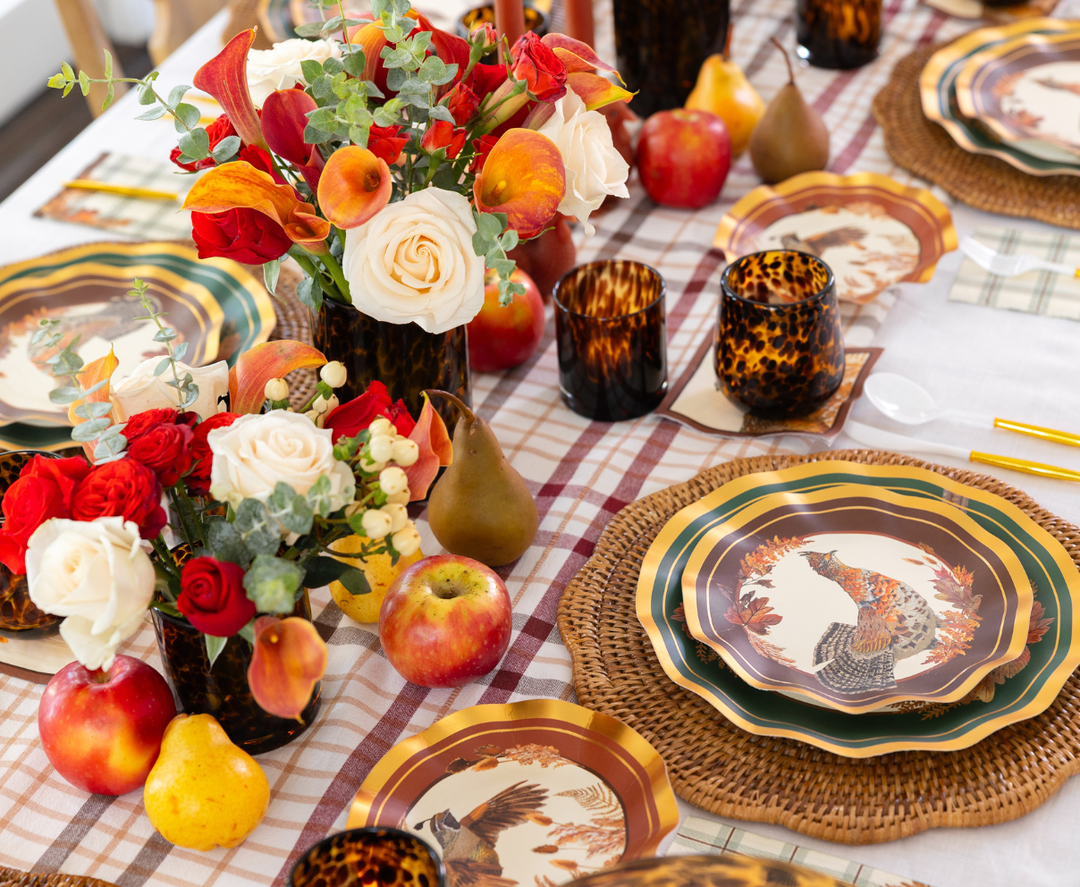 The Woodland Harvest Table Setting by Sophistiplate features autumn-themed designer paper plates, woven placemats, floral centerpieces, apples, pears, and tortoiseshell glasses on a plaid tablecloth—creating the perfect harvest party decor.