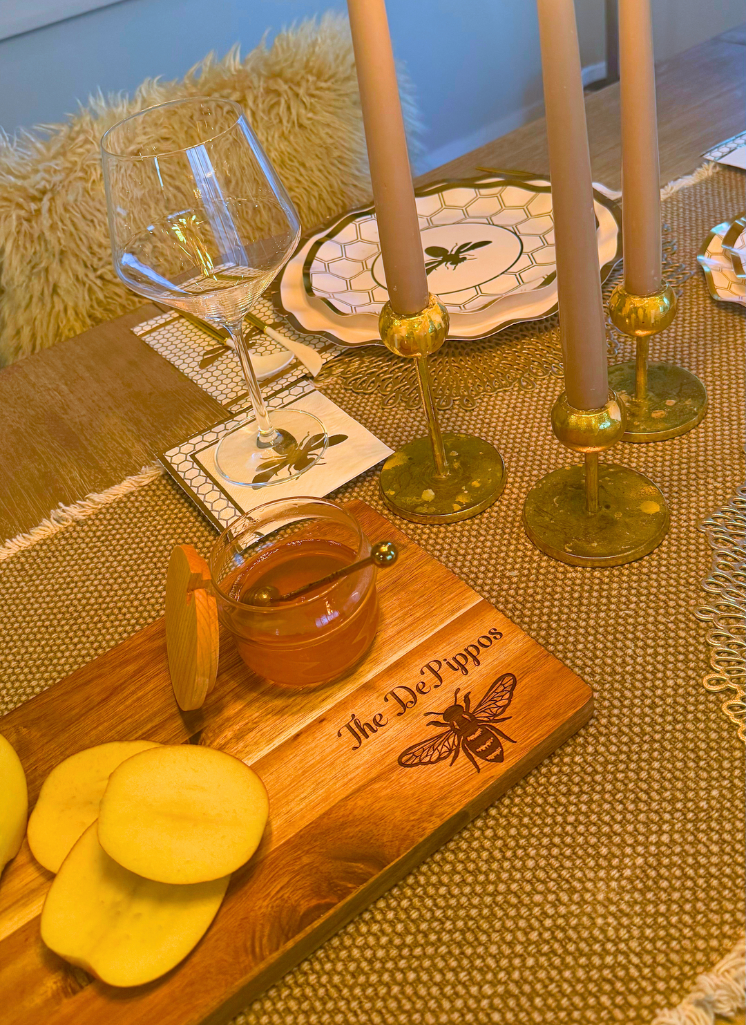 A 14″ x 8″ Acacia Wood Board by Maple, personalized with “The DePippos” and a charming honeybee illustration, showcases sliced apples and honey atop a table decorated with bee-themed plates, coasters, candles, and a wine glass. This unique board is perfect as a charcuterie or cheese board centerpiece for any gathering.