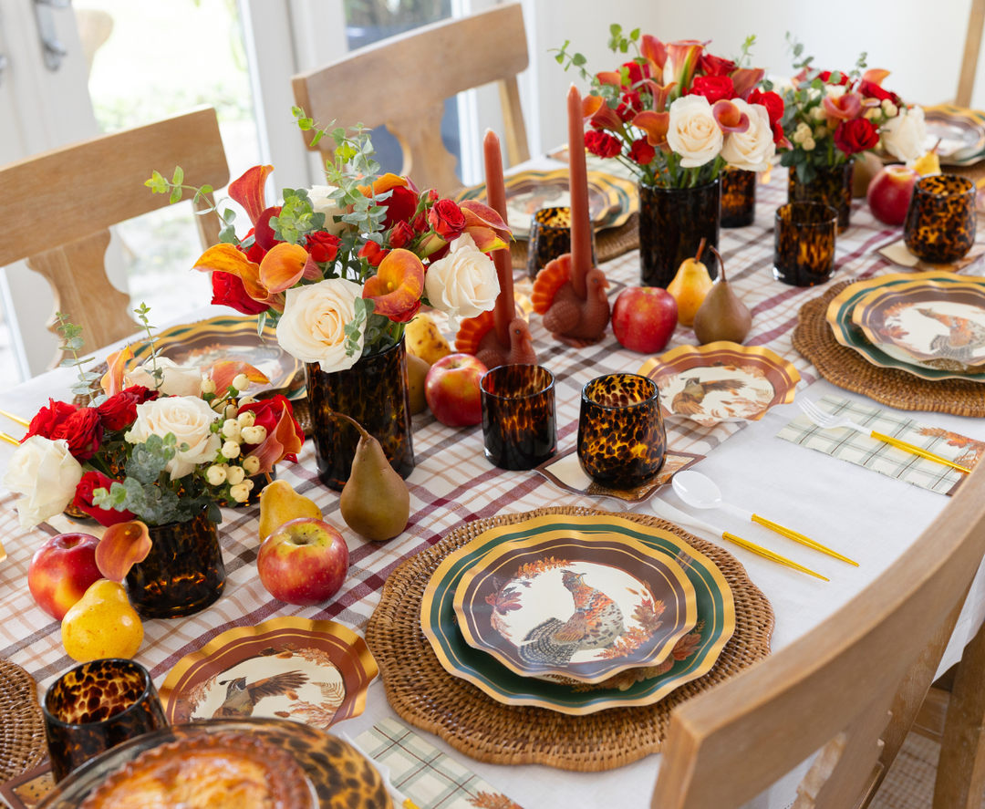 Create a festive harvest tablescape with the Sophistiplate Woodland Harvest Table Setting, featuring designer paper plates adorned with rooster motifs, tortoiseshell glasses, red and white floral centerpieces, turkey-shaped candles, and an arrangement of apples and pears atop a plaid tablecloth.