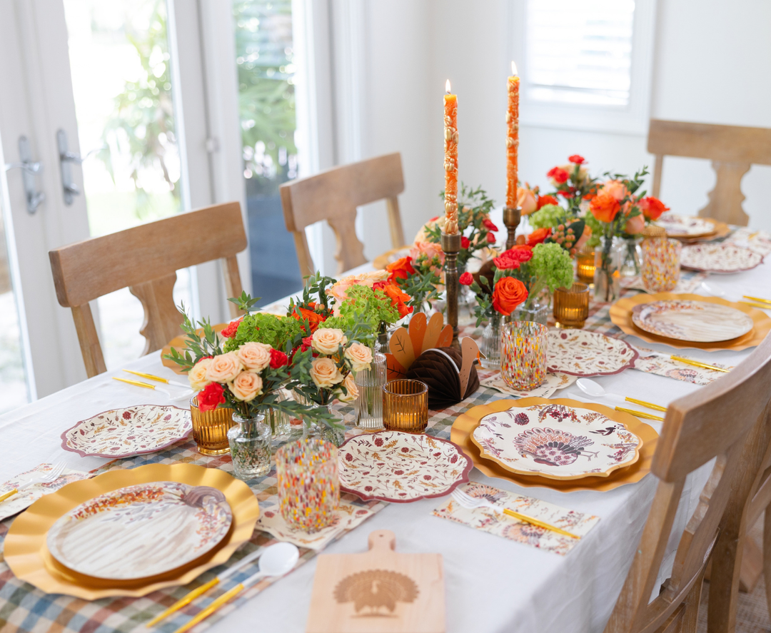 The Amber Harvest Table Setting by Sophistiplate creates a festive dining experience with floral arrangements, candles, patterned plates, and yellow utensils atop a white tablecloth. The autumn tablescape features orange and green accents, while Sophistiplate’s harvest party plates add seasonal charm to the setting.