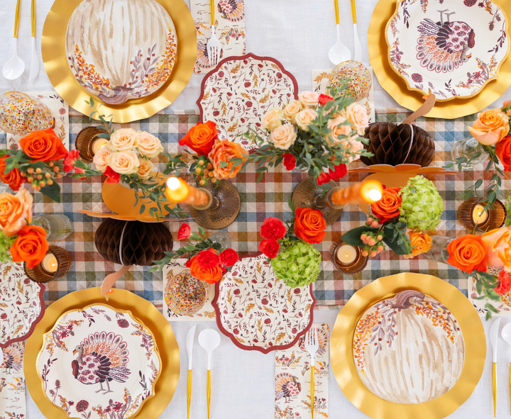 Create a festive autumn tablescape with the Amber Harvest Table Setting by Sophistiplate, showcasing rich seasonal colors. This collection features harvest party plates decorated with turkey and pumpkin motifs, complemented by gold chargers, orange and peach florals, warm candlelight, and a plaid table runner—perfect for a cozy fall celebration.