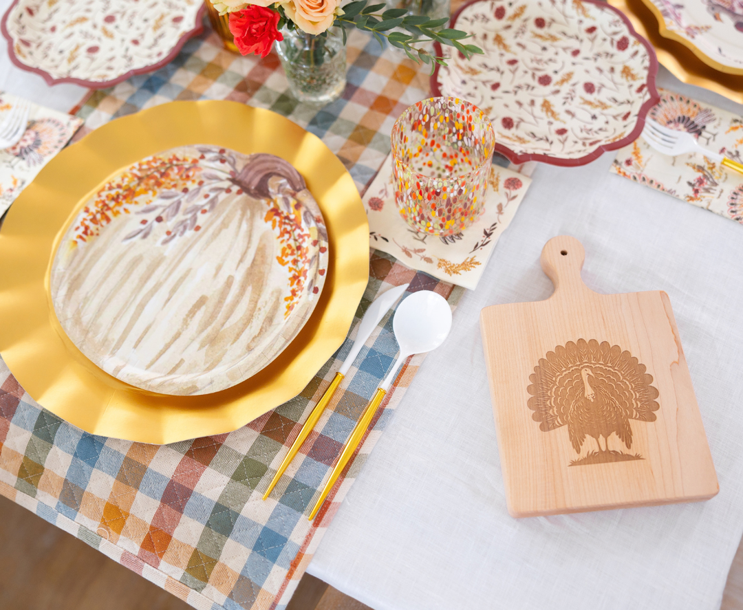The Amber Harvest Table Setting by Sophistiplate creates a festive autumn tablescape with harvest party plates layered on a gold charger, paired with a confetti dot glass and a wooden turkey-engraved cutting board, all styled on a plaid-and-white tablecloth with floral napkins and fresh flowers.