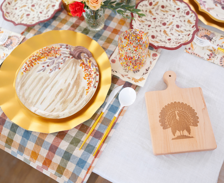 The Amber Harvest Table Setting by Sophistiplate creates a festive autumn tablescape with harvest party plates layered on a gold charger, paired with a confetti dot glass and a wooden turkey-engraved cutting board, all styled on a plaid-and-white tablecloth with floral napkins and fresh flowers.