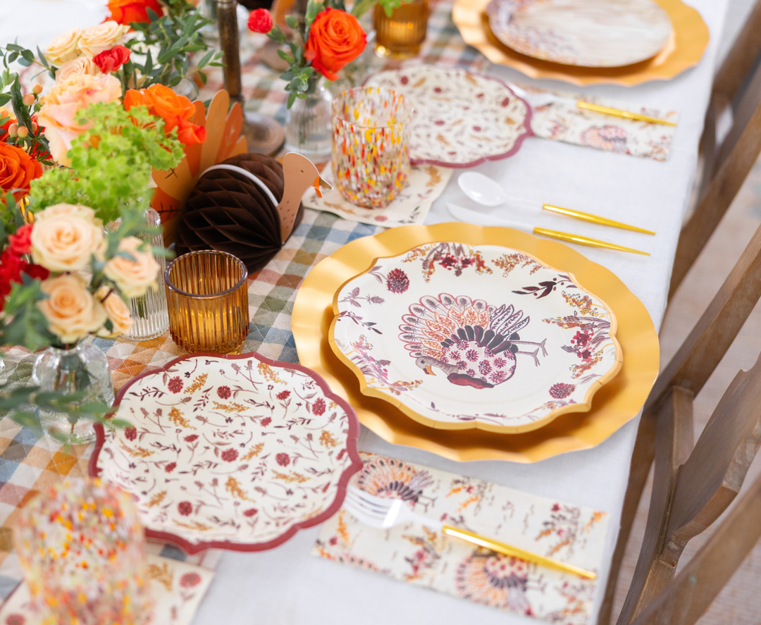 The Amber Harvest Table Setting by Sophistiplate creates a festive autumn tablescape with gold-edged disposable plates adorned with a turkey motif, complemented by autumn-themed napkins, orange flowers, candles, and a checkered tablecloth—perfect for a Thanksgiving celebration.