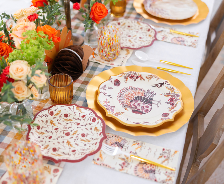 The Amber Harvest Table Setting by Sophistiplate creates a festive autumn tablescape with gold-edged disposable plates adorned with a turkey motif, complemented by autumn-themed napkins, orange flowers, candles, and a checkered tablecloth—perfect for a Thanksgiving celebration.