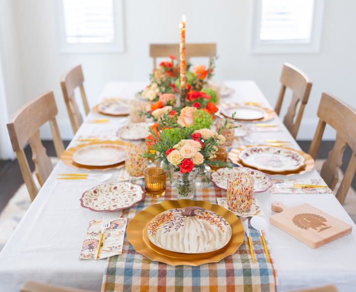 A festive autumn tablescape featuring the Sophistiplate Amber Harvest Table Setting is adorned with floral designer disposable plates, gold cutlery, a plaid runner, orange and peach flowers, and a glowing candle at its center. Wooden chairs encircle the table, ready for a meal.