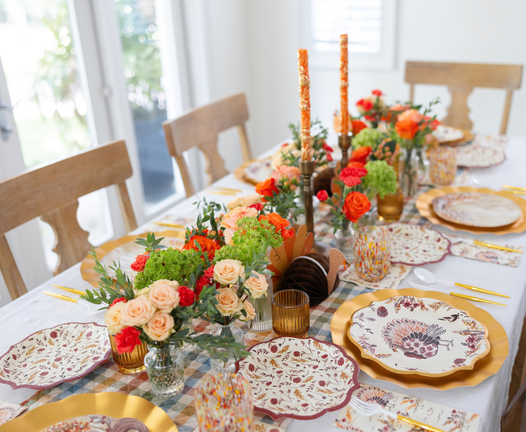 The Amber Harvest Table Setting by Sophistiplate creates a cheerful, elegant atmosphere with bright floral centerpieces, orange candles, and patterned turkey plates from the harvest party collection, beautifully complemented by gold chargers and glasses for a festive autumn tablescape.