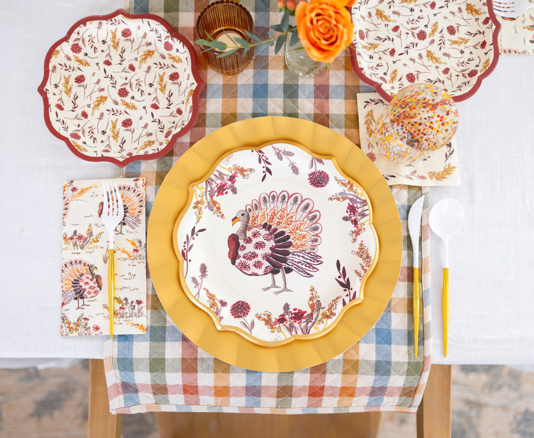 Elevate your festive harvest meal or gathering with the Amber Harvest Table Setting by Sophistiplate. This colorful autumn tablescape features turkey-themed designer disposable plates, a yellow charger, floral napkins, and plastic flatware, all beautifully arranged alongside an orange flower centerpiece on a plaid runner and crisp white tablecloth.