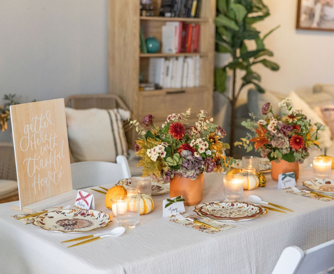 A cozy living room is beautifully decorated for a fall-themed meal, featuring the Sophistiplate Amber Harvest Table Setting with elegant designer disposable plates and festive harvest party plates. The tablescape includes floral centerpieces, candles, place cards, pumpkins, and a charming sign reading “Gather here with thankful hearts,” adding both charm and convenience to the autumn celebration.