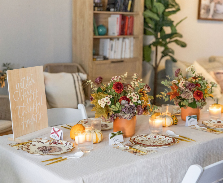 A cozy living room is beautifully decorated for a fall-themed meal, featuring the Sophistiplate Amber Harvest Table Setting with elegant designer disposable plates and festive harvest party plates. The tablescape includes floral centerpieces, candles, place cards, pumpkins, and a charming sign reading “Gather here with thankful hearts,” adding both charm and convenience to the autumn celebration.