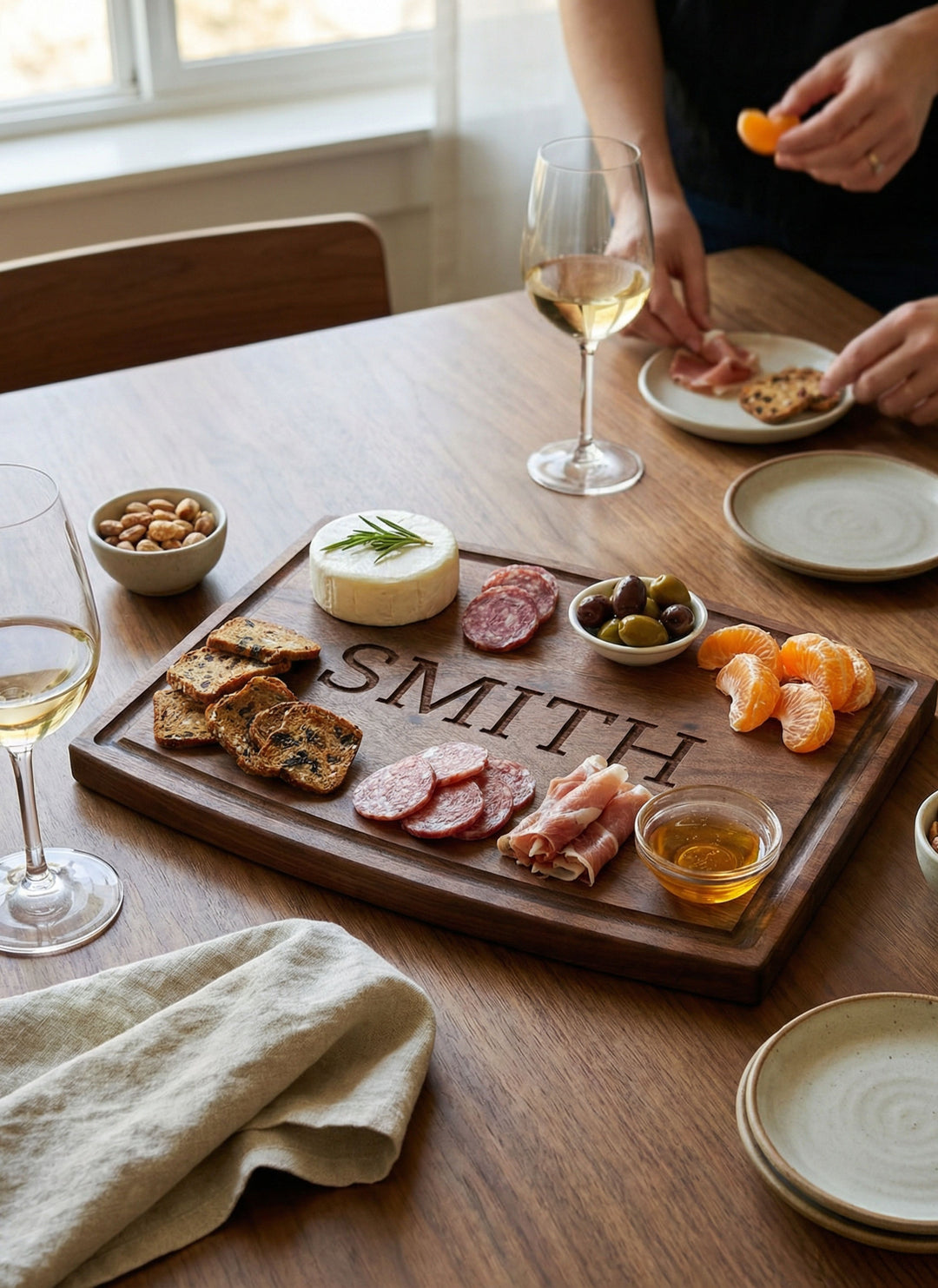 A Maple Personalized Walnut Wood Cutting + Cheeseboard (12 x 9") labeled SMITH displays cheese, crackers, salami, prosciutto, olives, nuts, honey, and tangerine slices on a wooden table set with two wine glasses and plates.