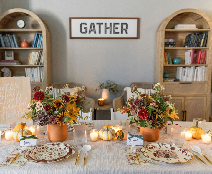 An inviting dining table is elegantly arranged with the Amber Harvest Table Setting by Sophistiplate, showcasing a stunning autumn display of floral centerpieces, candles, and festive harvest party plates. Bookshelves and a “GATHER” sign provide a charming backdrop, while two chairs await, promising a stylish celebration of the season.