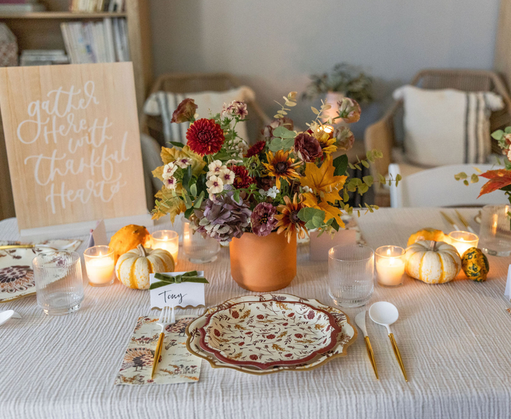 A festive autumn tablescape featuring the Sophistiplate Amber Harvest Table Setting is beautifully adorned with autumn flowers, candles, small pumpkins, and a sign that reads, Gather Here with Thankful Hearts. Plates, utensils, glasses, and a personalized name card for Tony are arranged neatly for a warm and inviting gathering.