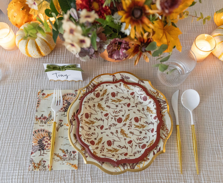 Create a fall-inspired tablescape with the Amber Harvest Table Setting by Sophistiplate, featuring floral-patterned harvest party plates, elegant gold and white flatware, an autumn-themed napkin, a coordinating name card, glowing candles, mini pumpkins, and a vibrant vase of seasonal flowers.