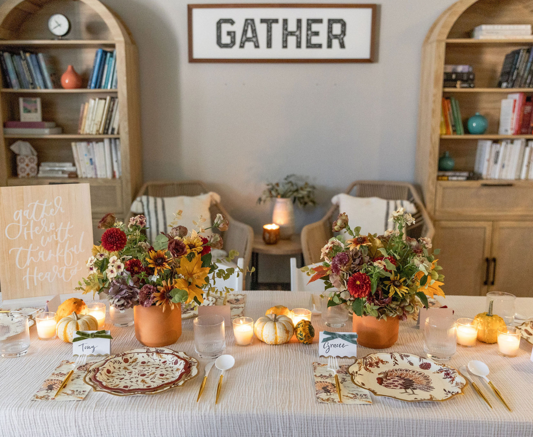 Experience the warmth of autumn with the Amber Harvest Table Setting by Sophistiplate. The dining table is beautifully arranged for fall, featuring flowers, candles, petite pumpkins, and personalized place cards. In the background, bookshelves and a GATHER sign enhance the inviting ambiance. Designer disposable plates from Sophistiplate’s Amber Harvest collection provide an elegant touch and effortless convenience to this charming seasonal scene.