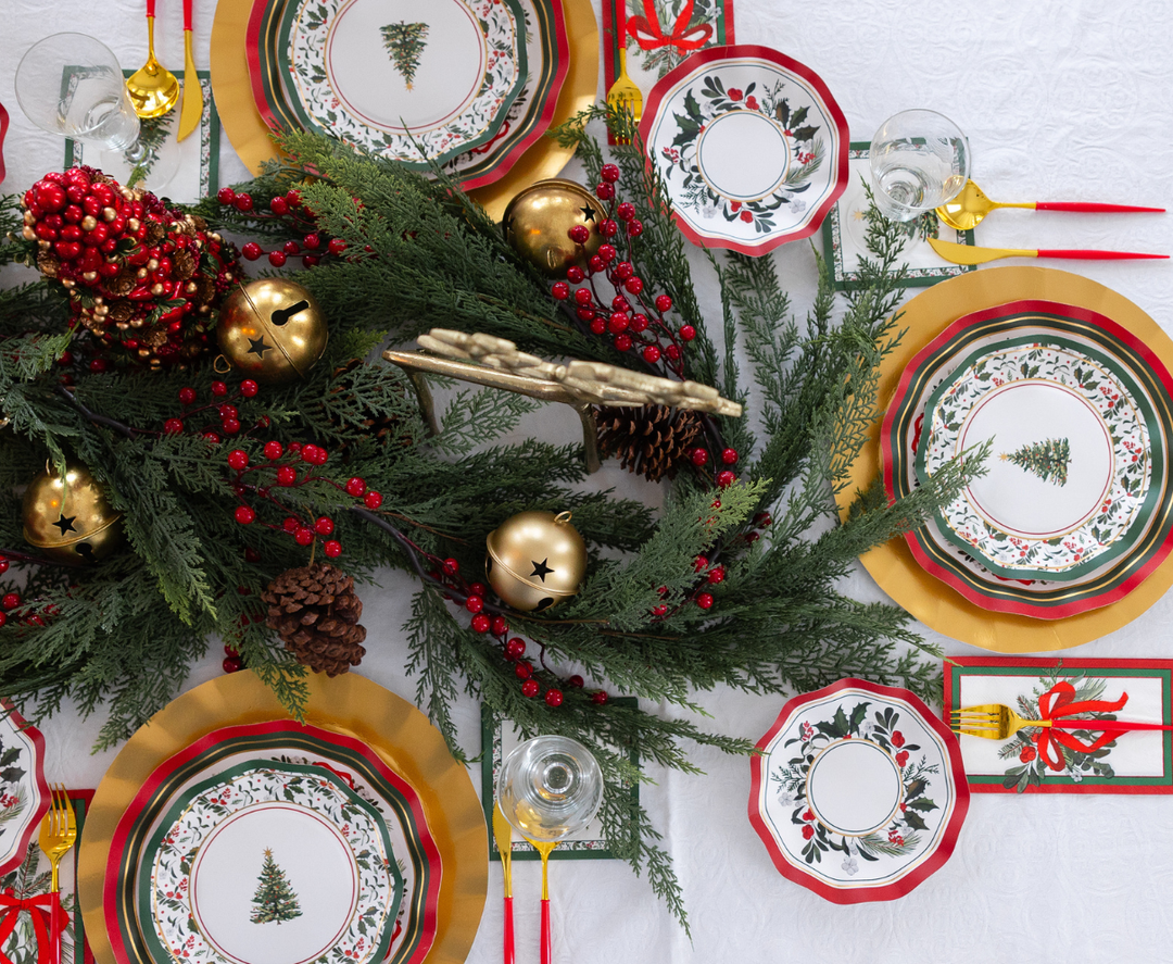 A Sophistiplate LLC Home for the Holidays: Christmas Table for 16 setting features heavy-duty paper plates with tree and berry designs, gold chargers, and red-handled cutlery around a centerpiece of evergreen branches, pinecones, and gold bells.
