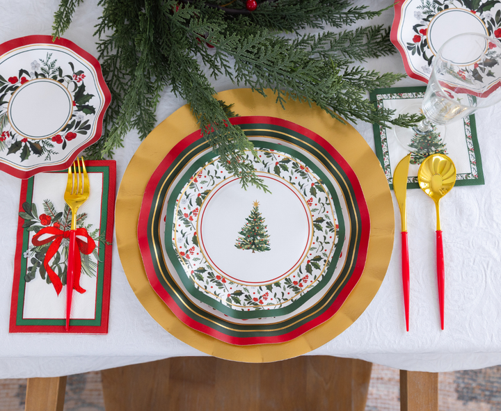 Home for the Holidays: Christmas Table for 16 setting featuring a white, heavy-duty paper plate with a Christmas tree design, a gold charger, a gold fork and spoon with red handles, and a napkin decorated with pine branches and a red ribbon.