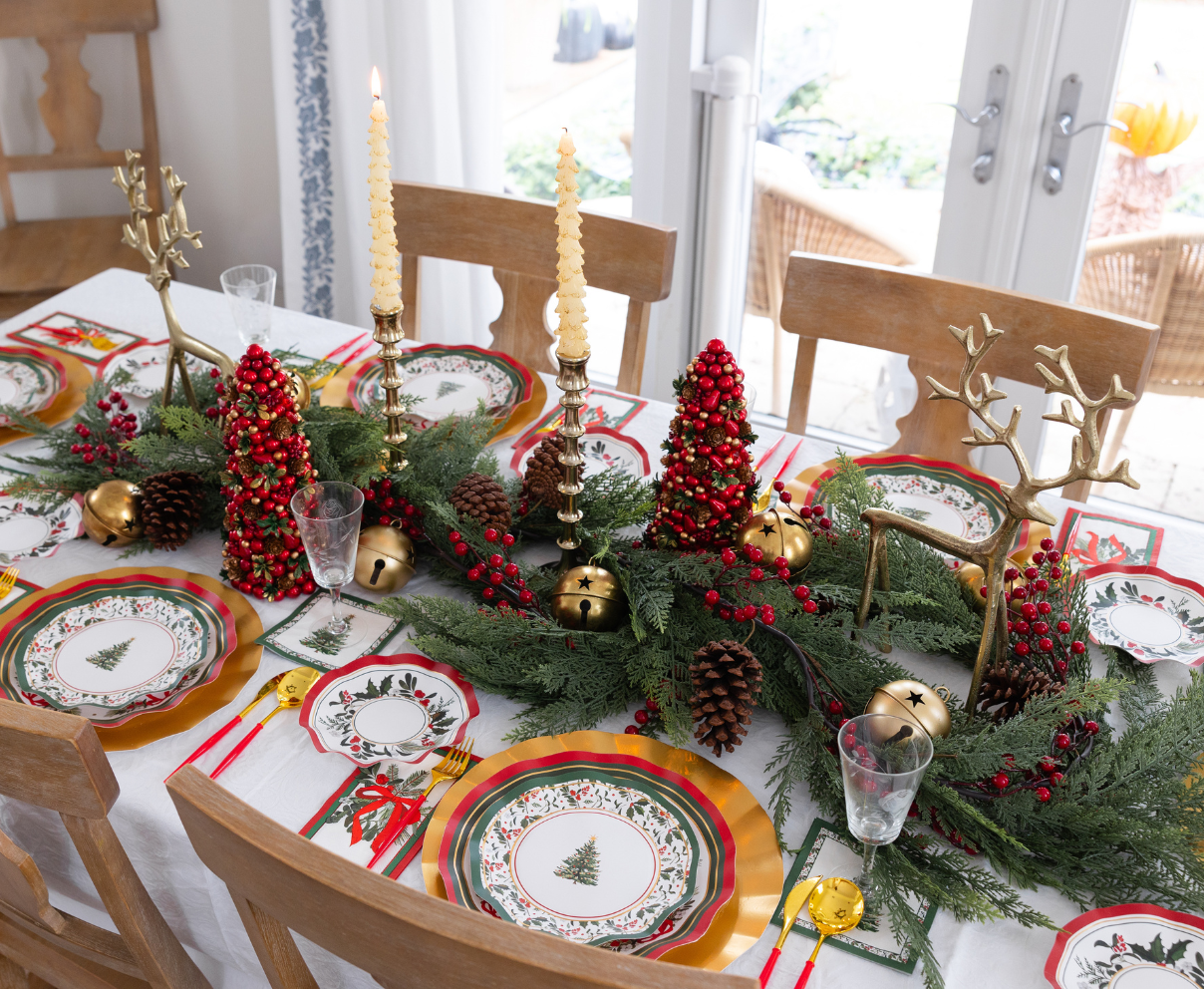 A Home for the Holidays: Christmas Table for 16 setting features heavy-duty paper plates with Christmas tree patterns, gold reindeer figures, red berry-covered miniature trees, and gold-colored cutlery on a white tablecloth.