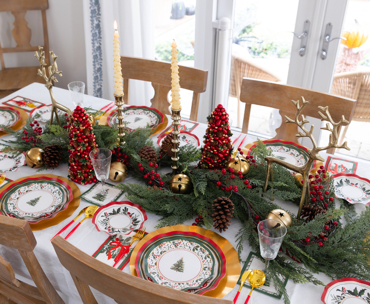 A Home for the Holidays: Christmas Table for 16 setting features heavy-duty paper plates with Christmas tree patterns, gold reindeer figures, red berry-covered miniature trees, and gold-colored cutlery on a white tablecloth.