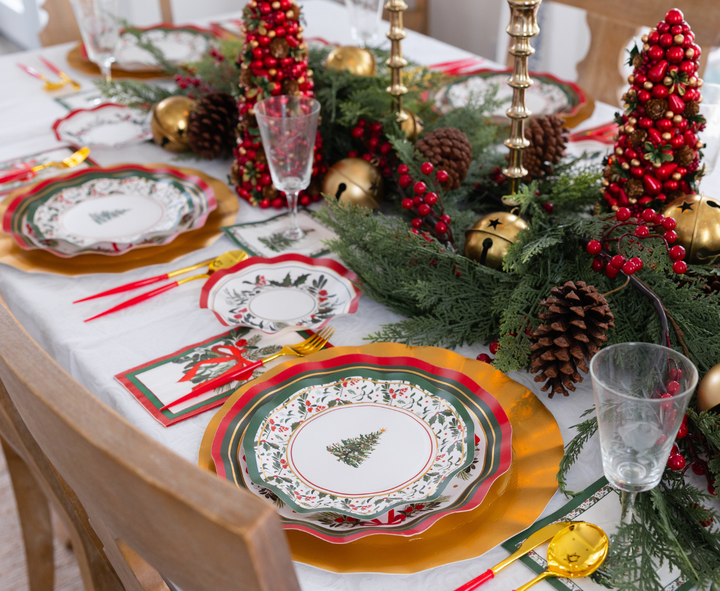 A Home for the Holidays: Christmas Table for 16 set features heavy duty paper plates with gold rims and tree illustrations, accented by red and gold ornaments, pine greenery, and gold cutlery.