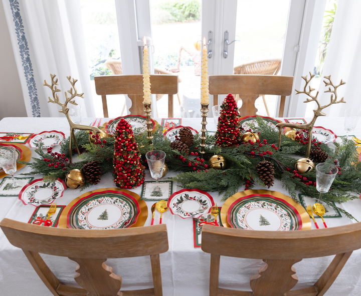 A Home for the Holidays: Christmas Table for 16 set with a centerpiece of pine branches, red beaded cone trees, gold reindeer figurines, and candles, featuring holiday-themed paper plates and gold-toned flatware.