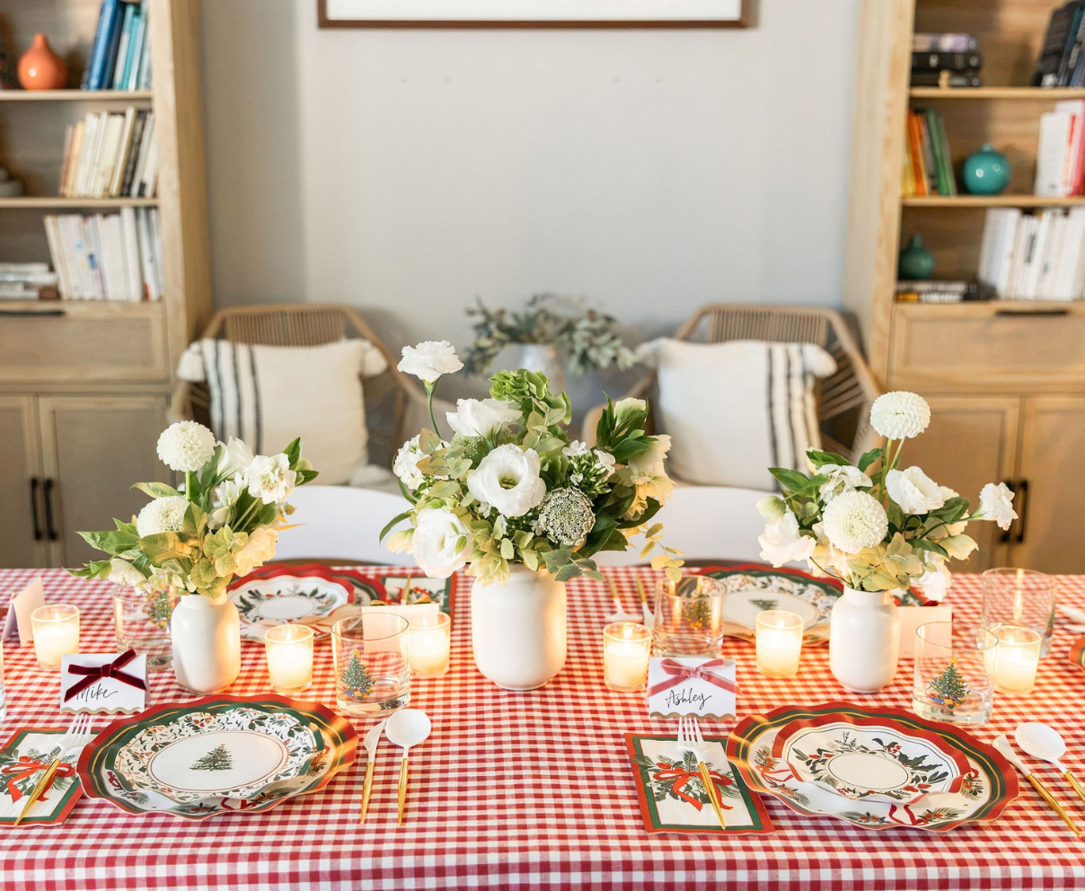 A Home for the Holidays: Christmas Table for 16 set for four with a red and white checkered tablecloth, Christmas-themed heavy duty paper plates, white floral centerpieces, and lit votive candles.