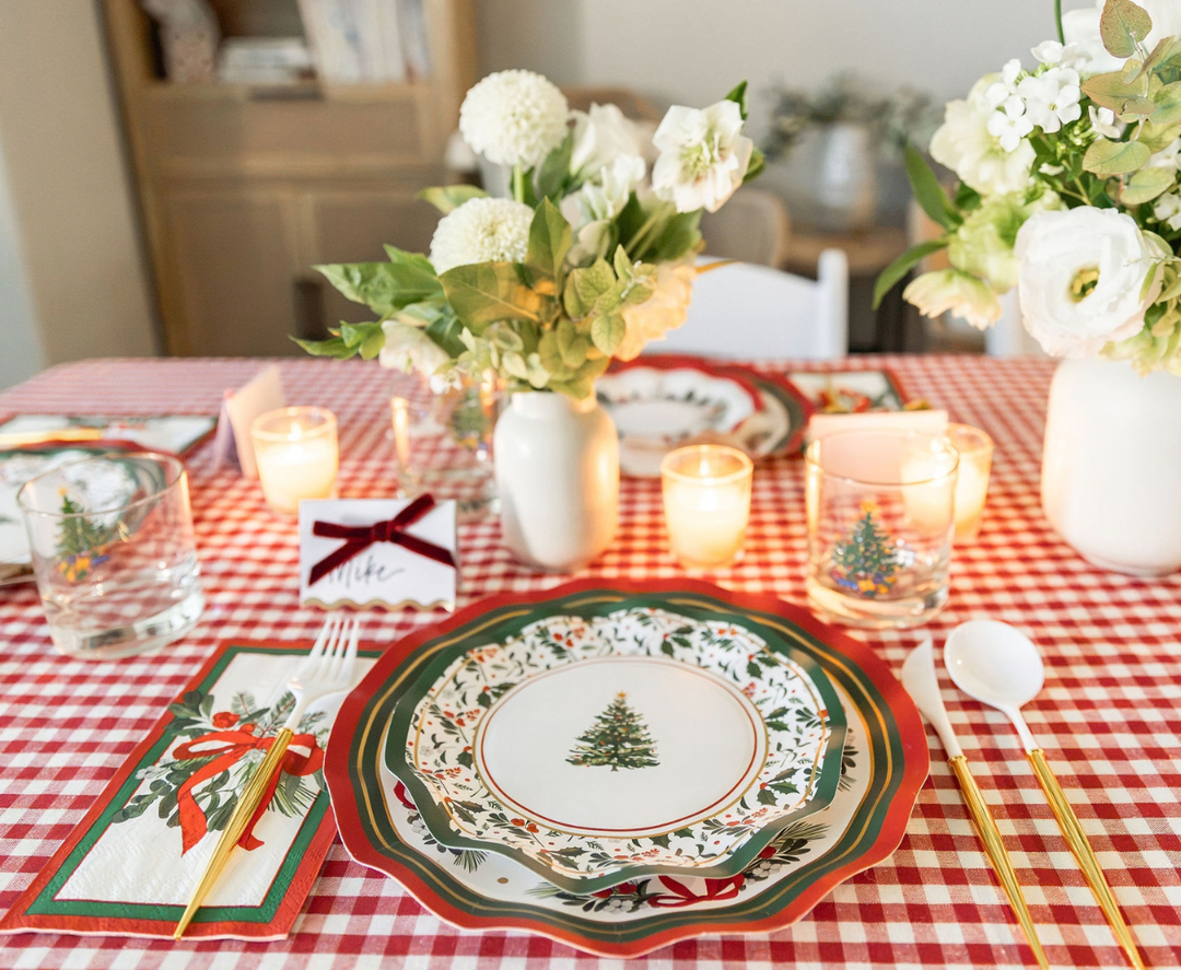 Home for the Holidays: Christmas Table for 16 set on a red and white checkered tablecloth, featuring layered heavy duty paper plates with a tree design, gold-handled cutlery, patterned napkins, and candles.