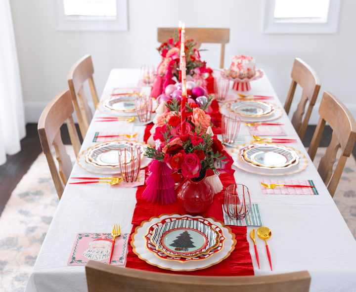 A Tis the Season: Christmas Table for 16 set for sixteen, featuring festive designer paper plates, red table runners, floral centerpieces, tall red candles, and Christmas napkins featuring Santa.