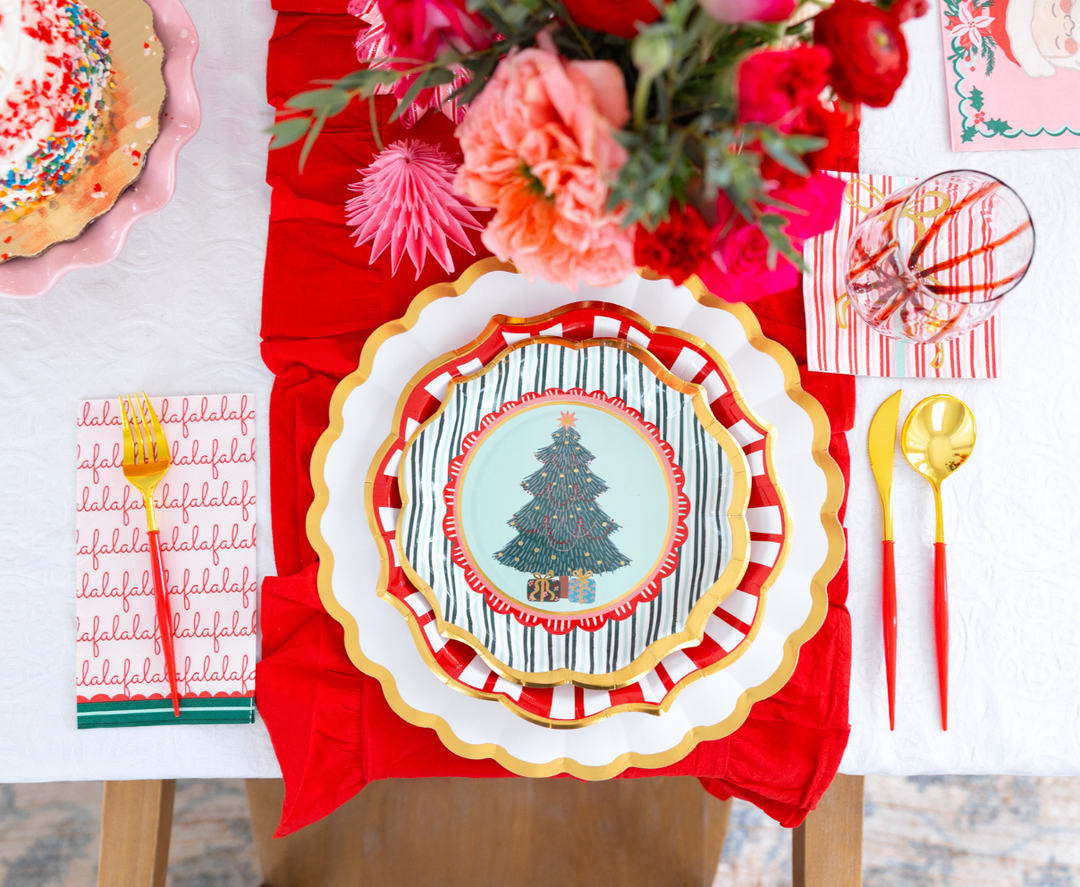 A Tis the Season: Christmas Table for 16 setting featuring a designer paper plate with a decorated tree, red and white striped salad plate, gold-rimmed white charger, red flatware, and a napkin printed with the word falalalala.