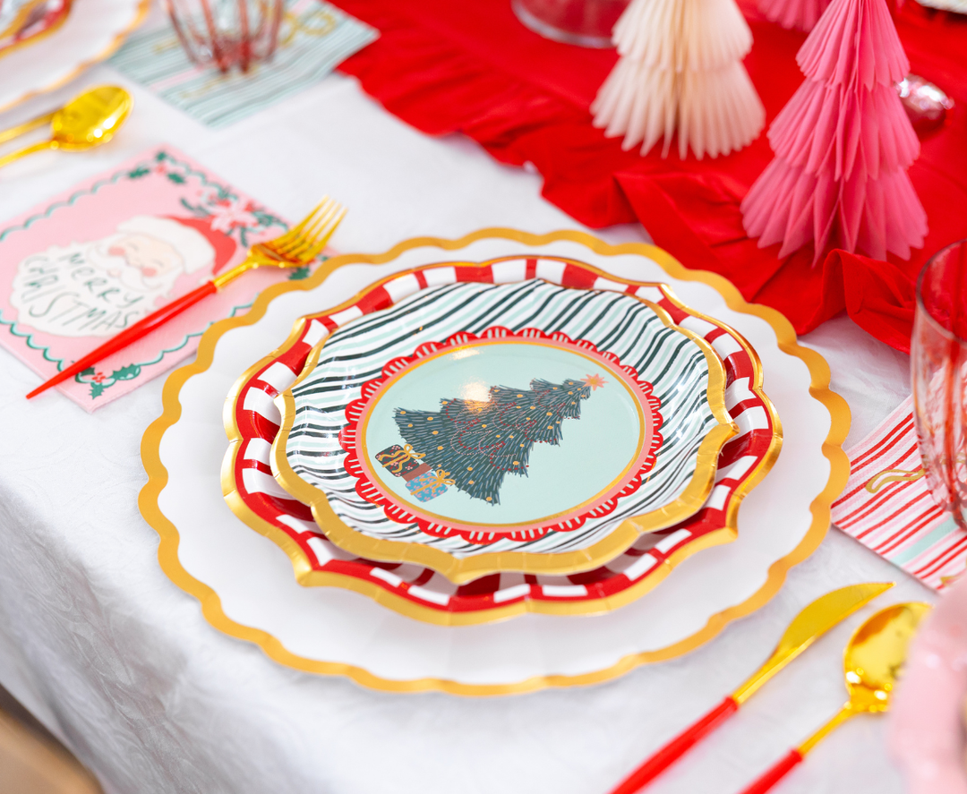 A Tis the Season: Christmas Table for 16 place setting featuring a scalloped charger, a red and white striped plate, a Christmas tree plate, gold-toned flatware with red handles, and a Santa Claus napkin for a festive Christmas table.