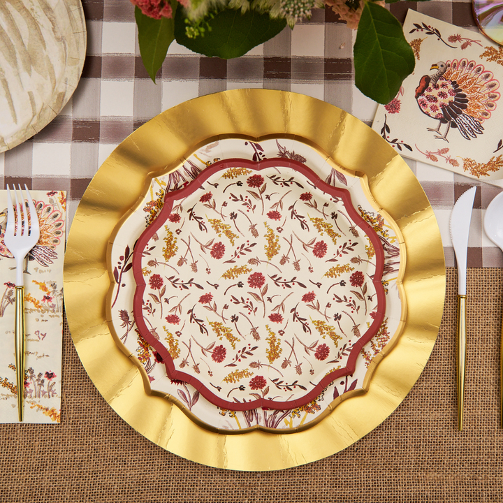 A decorative place setting featuring a gold charger, floral-patterned plate, white utensils with gold handles, napkins, and a bird-adorned paper accent—all arranged on a plaid and burlap tablecloth—creates an inviting tablescape. For added elegance and convenience, incorporate the Sophistiplate Amber Harvest Salad Plates (8-pack) as stylish disposable Thanksgiving plates to complete your festive look without compromising on style.