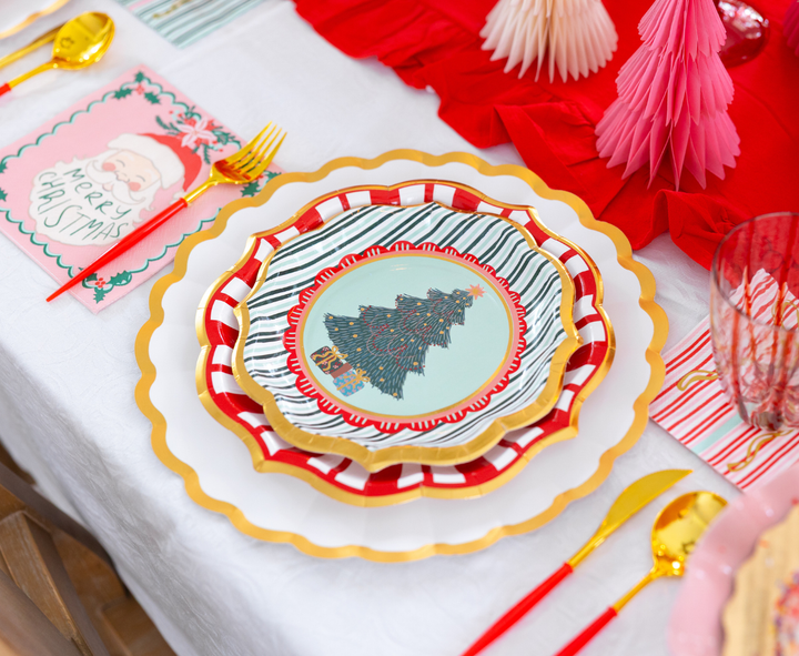 A Tis the Season: Christmas Table for 16 setting with layered scalloped designer paper plates featuring a tree design, a Santa-themed napkin, and gold-toned flatware with red handles.
