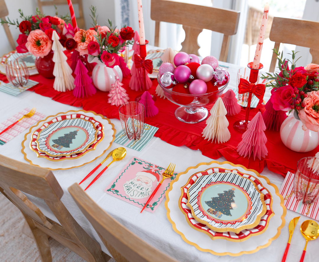 A Tis the Season: Christmas Table for 16 featuring designer paper plates with red and pink tree designs, matching napkins depicting Santa Claus, and a centerpiece of red flowers and pink ornaments.