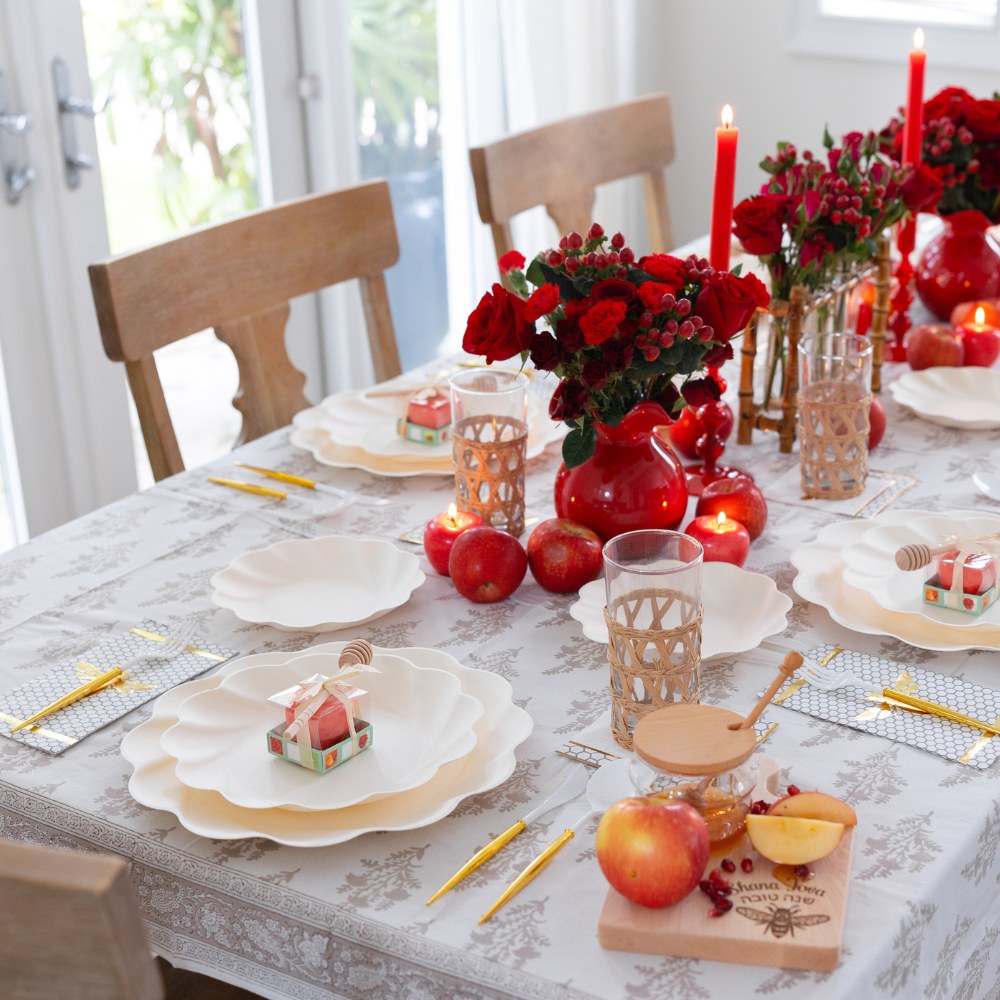 Elevate your special gathering with the Sophistiplate LLC “Rosh Tablescape for 16 Guests: Eco Cream Honeybee,” a beautiful Rosh Hashanah tableware set featuring elegant white plates, red candles, red roses in vases, apples, wicker glass holders, and charming mini gift boxes—all arranged on a sophisticated white-patterned tablecloth. Bathed in warm natural light, this setting creates a festive and inviting atmosphere perfect for celebrating with loved ones.