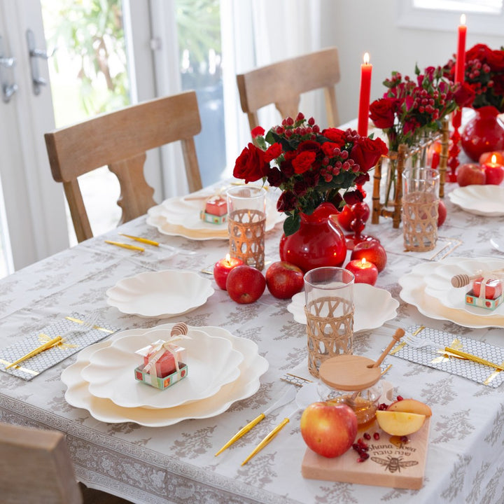 Elevate your special gathering with the Sophistiplate LLC “Rosh Tablescape for 16 Guests: Eco Cream Honeybee,” a beautiful Rosh Hashanah tableware set featuring elegant white plates, red candles, red roses in vases, apples, wicker glass holders, and charming mini gift boxes—all arranged on a sophisticated white-patterned tablecloth. Bathed in warm natural light, this setting creates a festive and inviting atmosphere perfect for celebrating with loved ones.