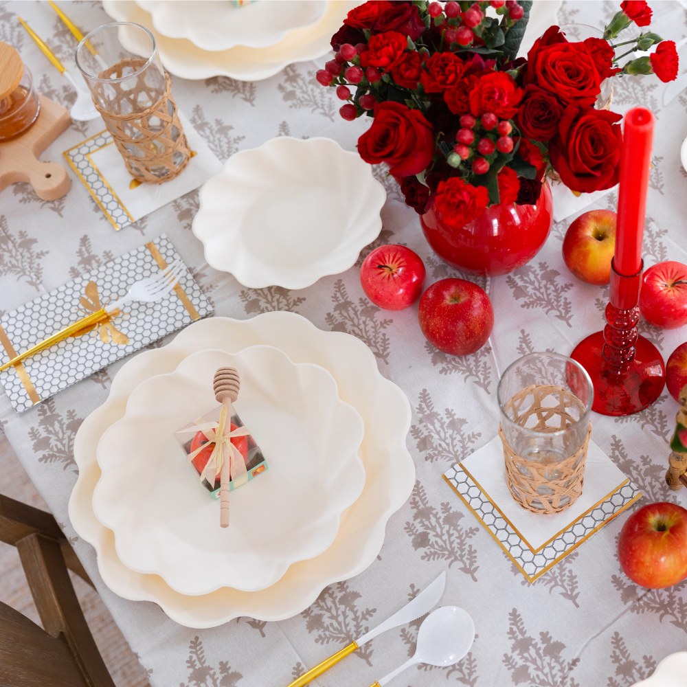 Elevate your celebration with the Sophistiplate LLC Rosh Tablescape for 16 Guests: Eco Cream Honeybee—a dazzling table set featuring scalloped white compostable plates, honey dippers paired with small jars, elegant glasses, red apples, a red vase of roses, and a glowing red candle atop a patterned tablecloth—perfectly blending stylish Rosh Hashanah tableware with eco-friendly convenience.