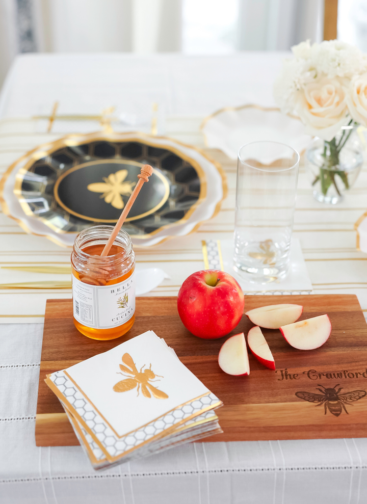 A table set with a bee-themed plate, honey jar with dipper, whole and sliced apple arranged on the Maple 14″ x 8″ Acacia Wood Board personalized with a honeybee, floral napkins, a glass of water, and a small bouquet of white roses.