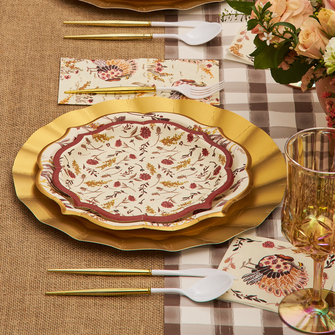 A table setting with Sophistiplate Amber Harvest Salad Plates (8pk), gold utensils, fancy-edge paper plates, plastic spoons, a plaid tablecloth, napkins with floral designs, and a bouquet of flowers in the corner.