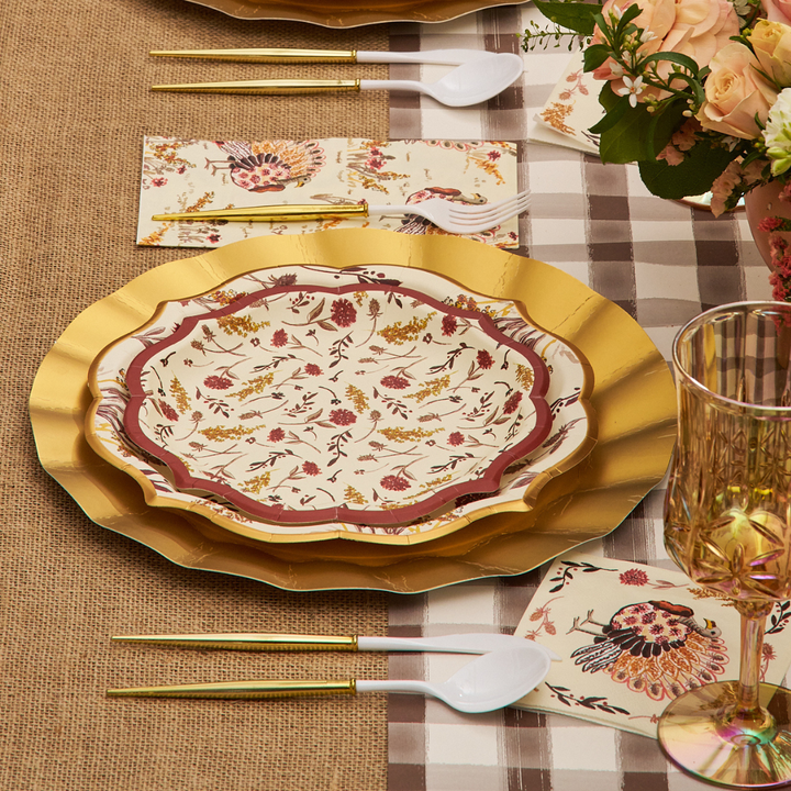 A table setting with Sophistiplate Amber Harvest Salad Plates (8pk), gold utensils, fancy-edge paper plates, plastic spoons, a plaid tablecloth, napkins with floral designs, and a bouquet of flowers in the corner.