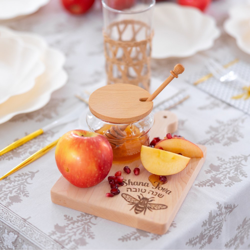 A wooden board adorned with an apple, peach slices, pomegranate seeds, and a honey jar sits gracefully atop the table. The board is inscribed with “Shana Tova” in both English and Hebrew, setting a festive tone for the occasion. In the background, Sophistiplate LLC’s Rosh Tablescape for 16 Guests: Eco Cream Honeybee—featuring compostable plates and utensils—rests on a crisp white tablecloth alongside glasses and plates, creating an inviting celebration atmosphere.