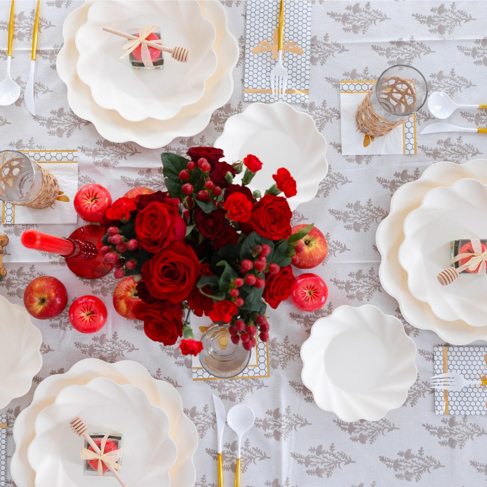 The Sophistiplate LLC “Rosh Tablescape for 16 Guests: Eco Cream Honeybee” features elegant cream scalloped plates, perfect as stylish disposable tableware for Rosh Hashanah or festive gatherings, complemented by gold cutlery, apples, red roses in a vase, glasses, and beautifully wrapped gifts displayed on a patterned tablecloth.