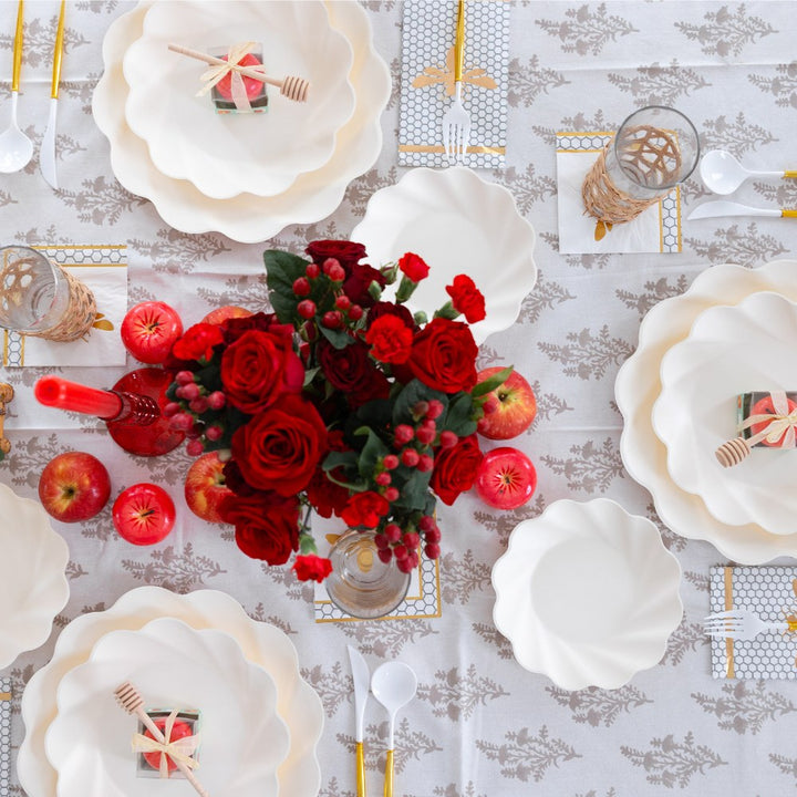 The Sophistiplate LLC “Rosh Tablescape for 16 Guests: Eco Cream Honeybee” features elegant cream scalloped plates, perfect as stylish disposable tableware for Rosh Hashanah or festive gatherings, complemented by gold cutlery, apples, red roses in a vase, glasses, and beautifully wrapped gifts displayed on a patterned tablecloth.
