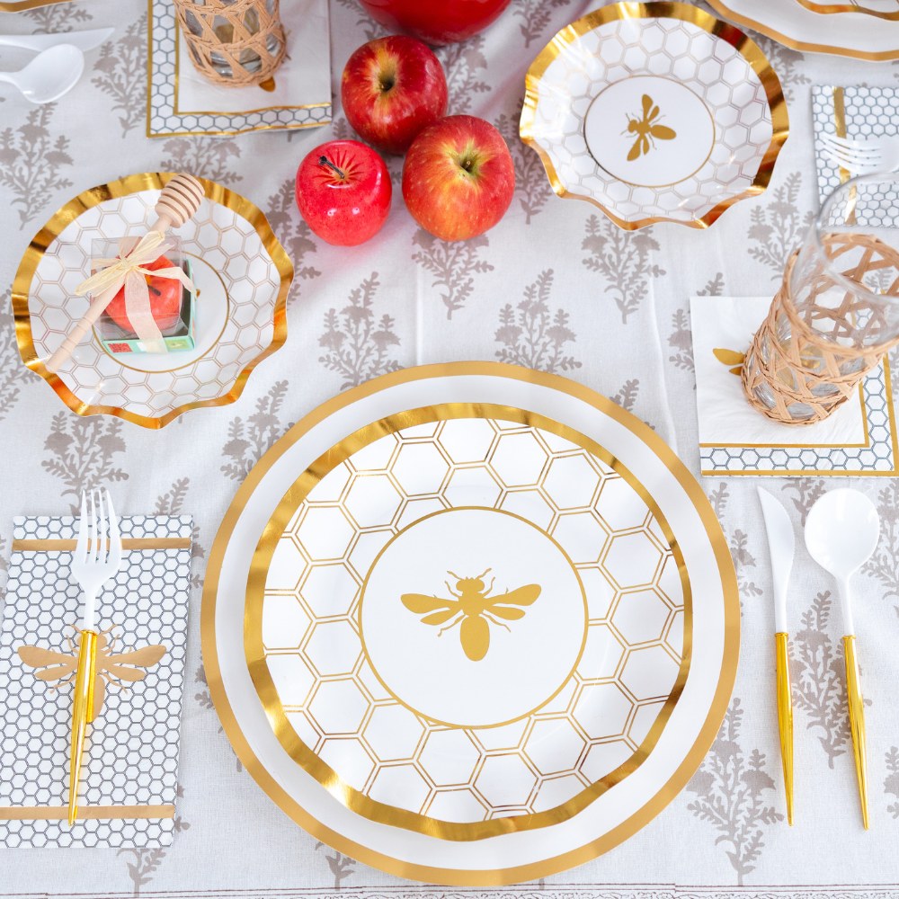 The table is set with the Sophistiplate White Honeybee Table Setting, featuring elegant disposable plates, bowls, and napkins with a gold and white bee motif. Three red apples and a pomegranate rest on the light floral-patterned tablecloth.
