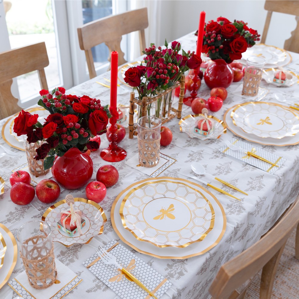 Create a festive Rosh Hashanah table with the Sophistiplate LLC “Rosh Tablescape for 16 Guests: White Honeybee” set, featuring designer disposable plates adorned with gold trim and bee motifs. Complete your elegant table setting with red flowers in vases, red candles, glassware, pomegranates, and a white-patterned tablecloth—perfect for celebrating the holiday in style.