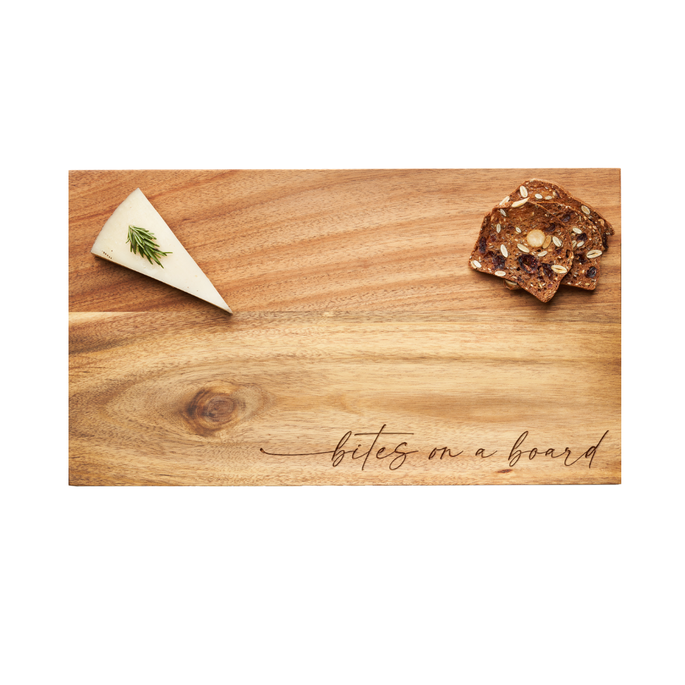 A wedge of herb-topped cheese and a stack of seeded crackers are served on the Maple Modern Acacia Rectangle board (14 x 8″), featuring the phrase “bites on a board” in elegant script at the bottom right—perfect for stylish entertaining.