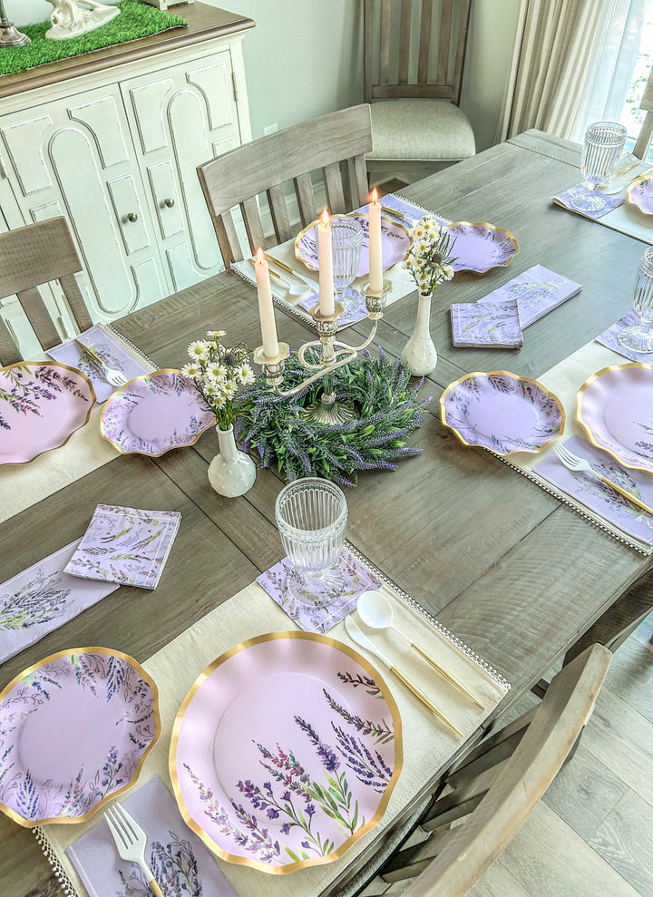 The Lavender Fields Table Setting by Sophistiplate LLC features a wooden dining table set for six with designer disposable plates, lavender-themed napkins, floral centerpieces, a candelabra with lit candles, and glassware on cream placemats.