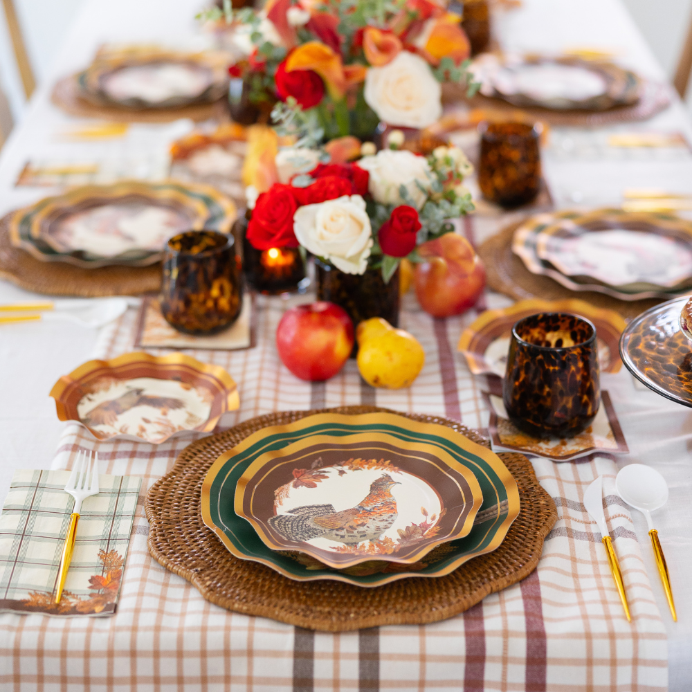 Create a vibrant autumn tablescape with the Woodland Harvest Table Setting by Sophistiplate, featuring designer paper plates adorned with pheasant motifs, woven placemats, plaid napkins, gold flatware, tortoise-shell cups, and a centerpiece of red, peach, and white flowers accented by apples and pears.