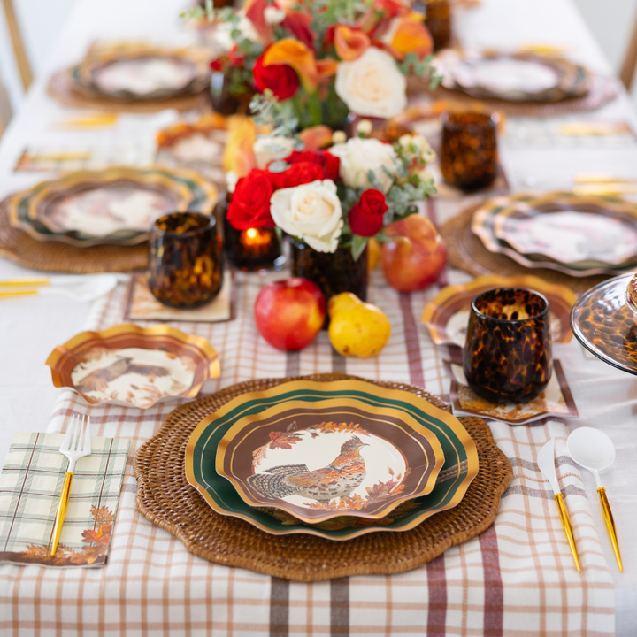 Create a vibrant autumn tablescape with the Woodland Harvest Table Setting by Sophistiplate, featuring designer paper plates adorned with pheasant motifs, woven placemats, plaid napkins, gold flatware, tortoise-shell cups, and a centerpiece of red, peach, and white flowers accented by apples and pears.