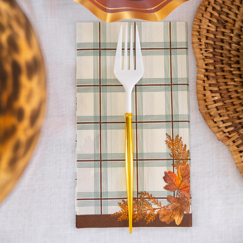 A white fork with a gold handle rests on a plaid napkin from the Sophistiplate Woodland Harvest Guest Towels, adorned with autumn leaves, set atop a white tablecloth beside woven placemats.